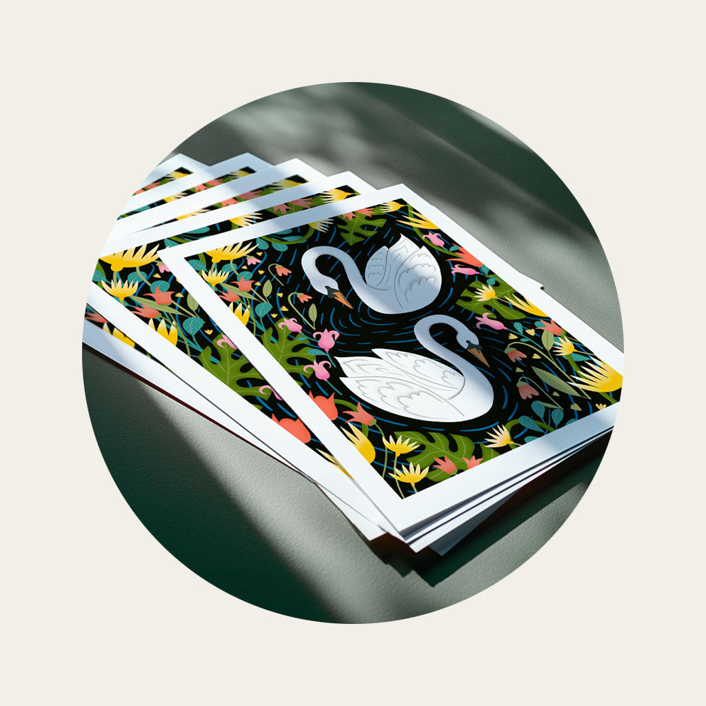 A stack of fine art prints on a tabletop, the artwork is an illustration of two swans surrounded by leaves and flowers.