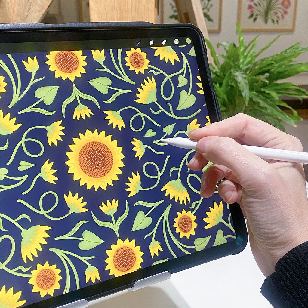 A hand uses a stylus to draw a digital sunflower pattern on a tablet screen.