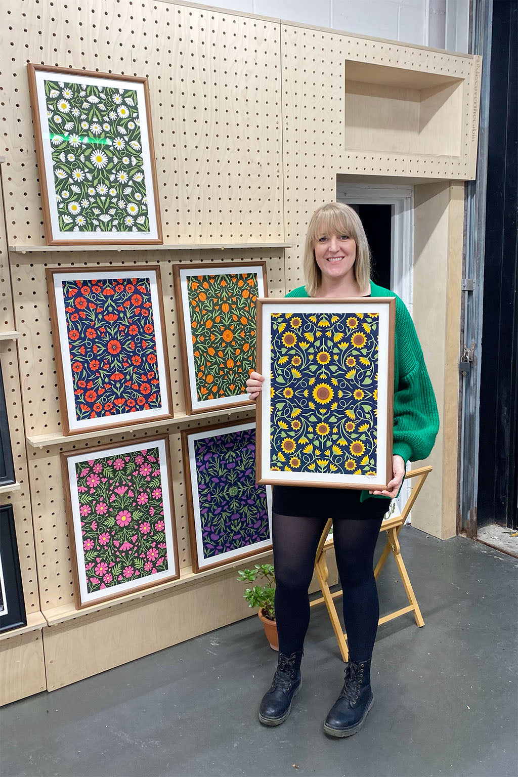 A woman in a green sweater holds a framed artwork with a floral pattern, standing by a display of similar framed art pieces on a pegboard wall.