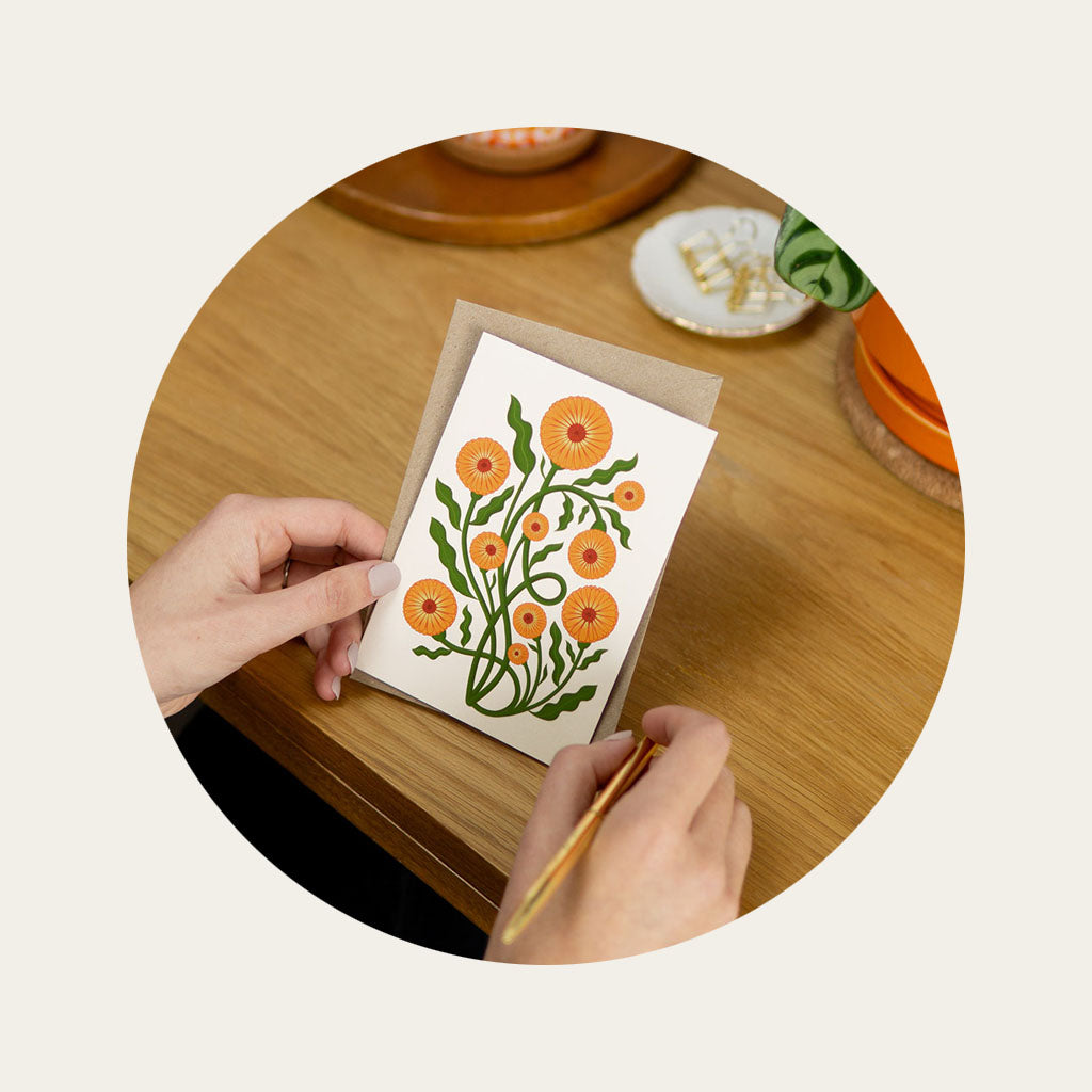 A woman's hand holding a greetings card that has a vintage style botanical illustration of a marigold with orange flowers.