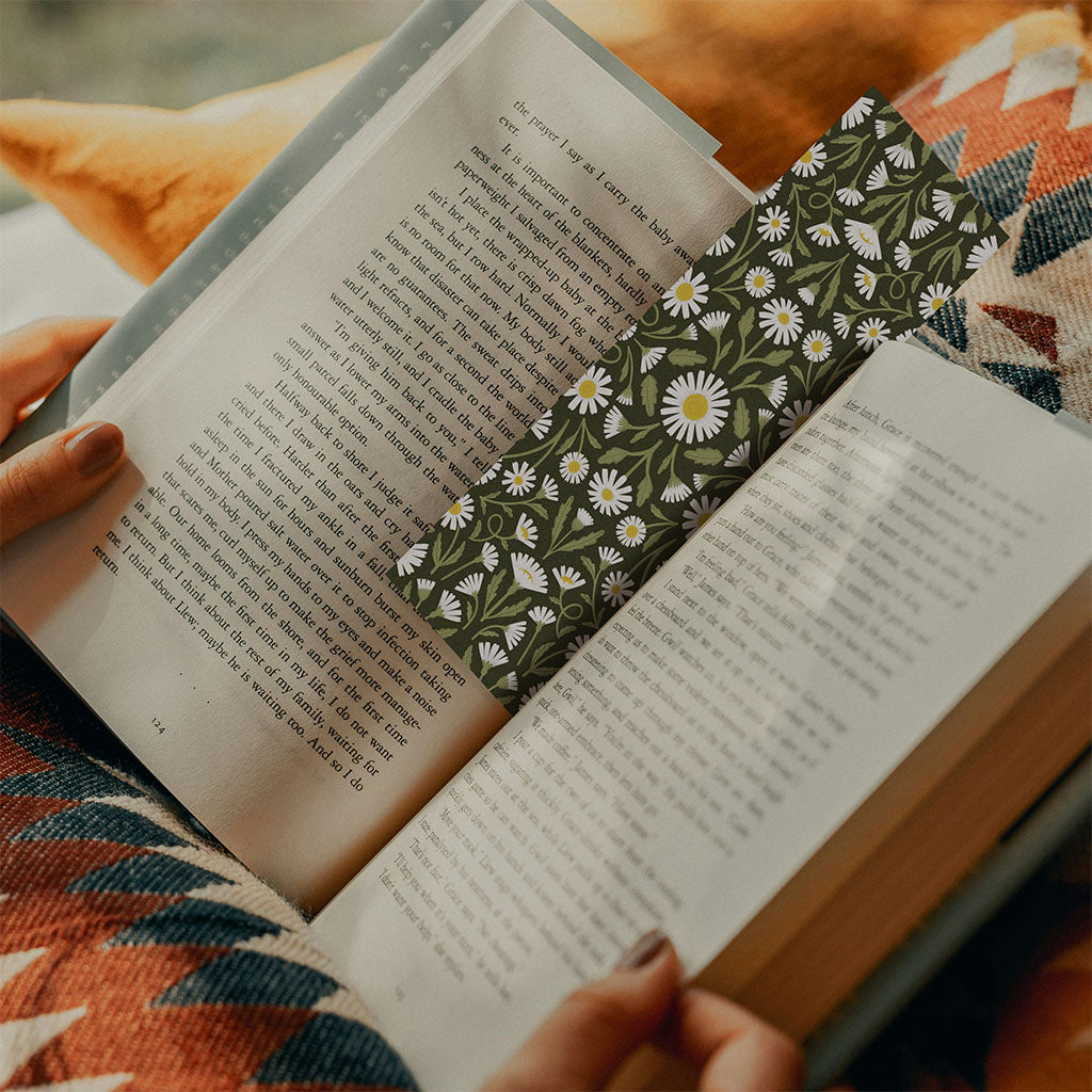 A person enjoying a cozy reading moment on a patterned blanket uses the Heritage Daisy Bookmark, adding vintage charm as it peeks from their open book.
