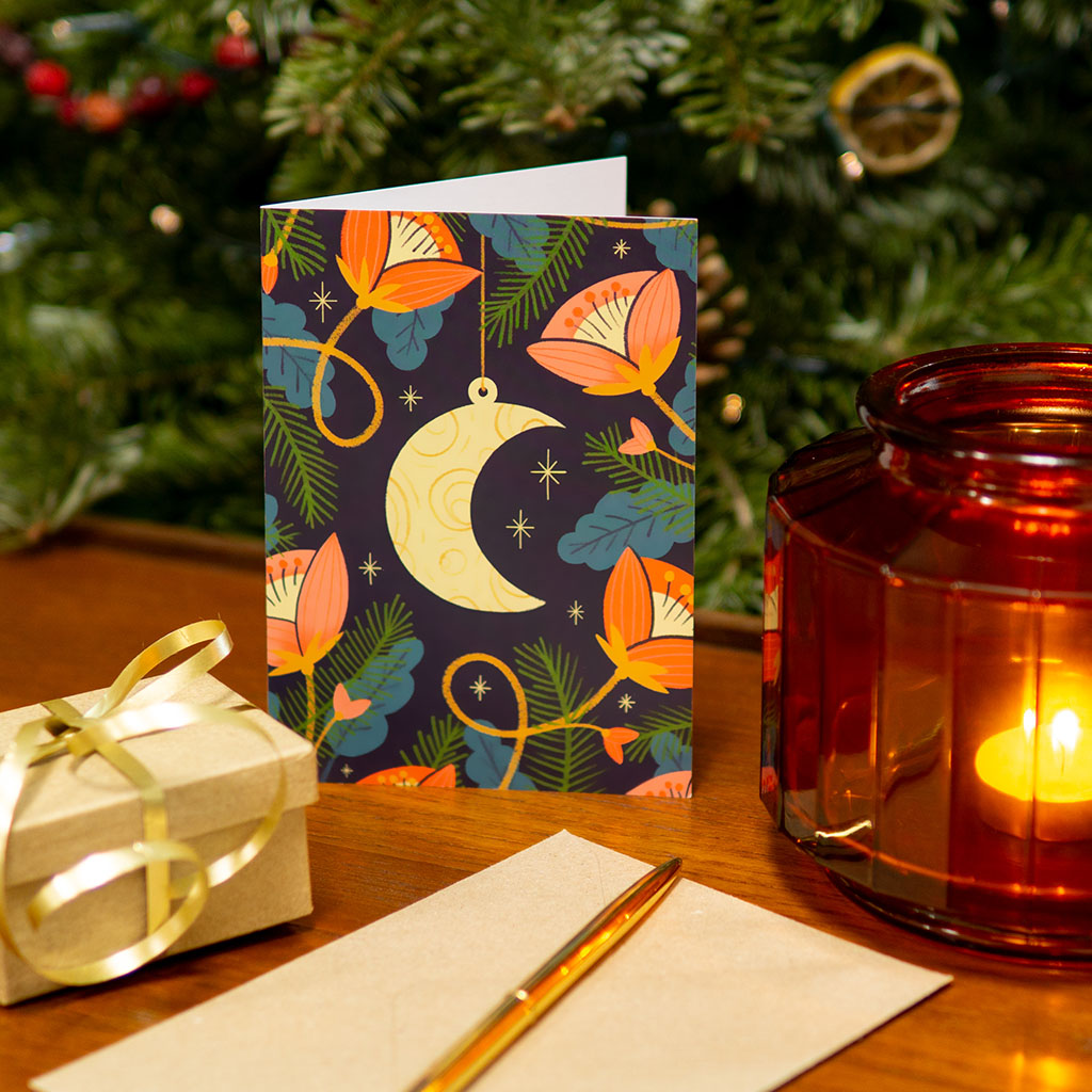 The Moon Christmas Card features a gold crescent moon pendant surrounded by pink flowers, green leaves, and small stars on a dark background, adding holiday magic.