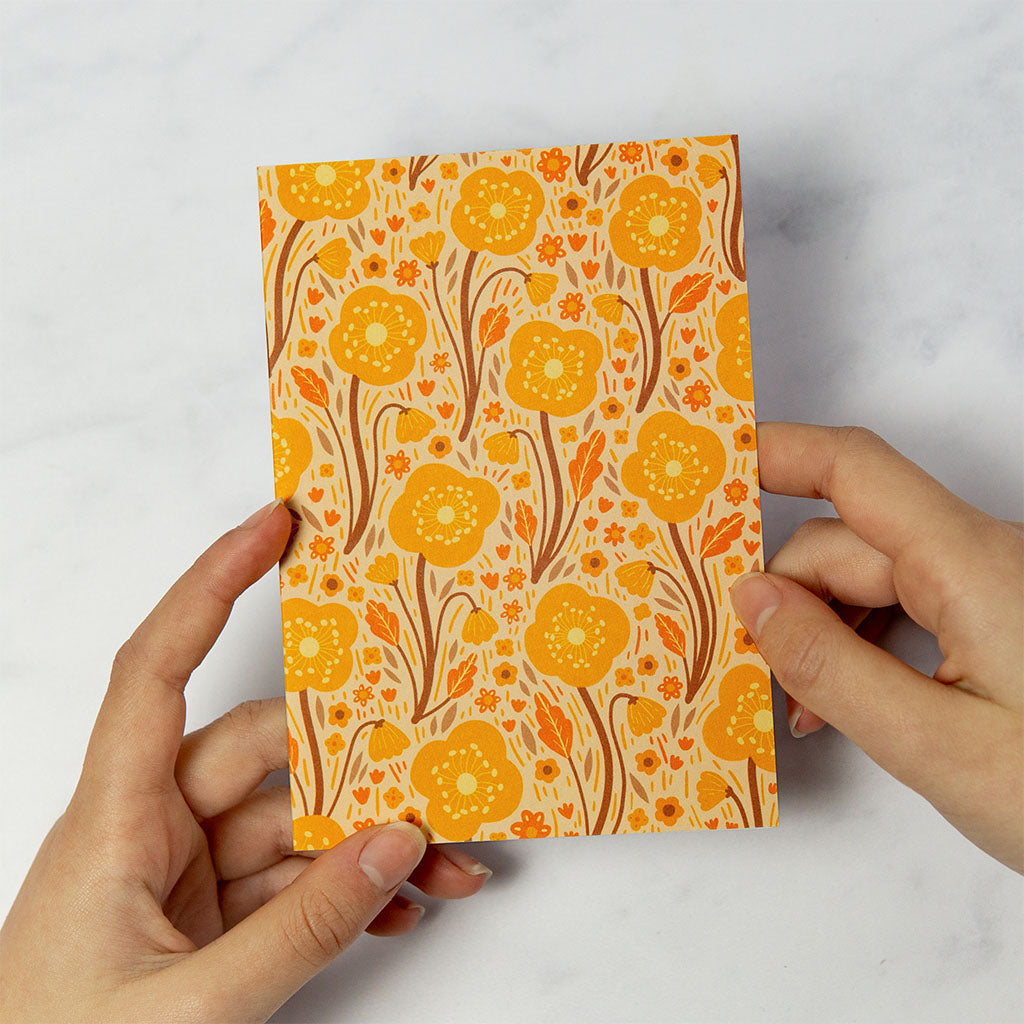 The Orange Wildflowers Greetings Card stands upright, showcasing a yellow and orange floral design with illustrated botanical British wildflowers and leaves on a beige background.