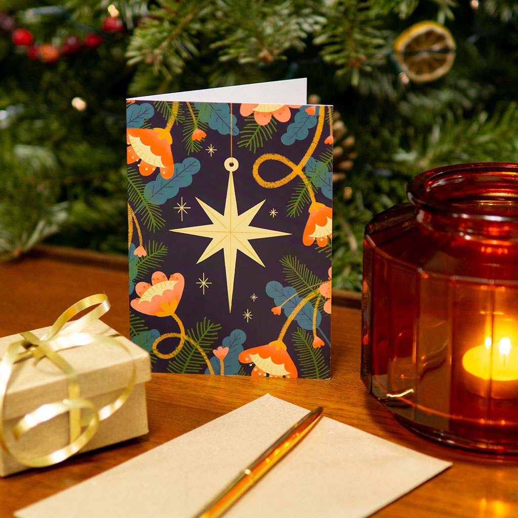 The Star Christmas Card features a gold star ornament with red flowers, green leaves, and pine branches on a dark background. Printed with vegetable-based inks on sustainable materials, each card comes with a kraft paper envelope.
