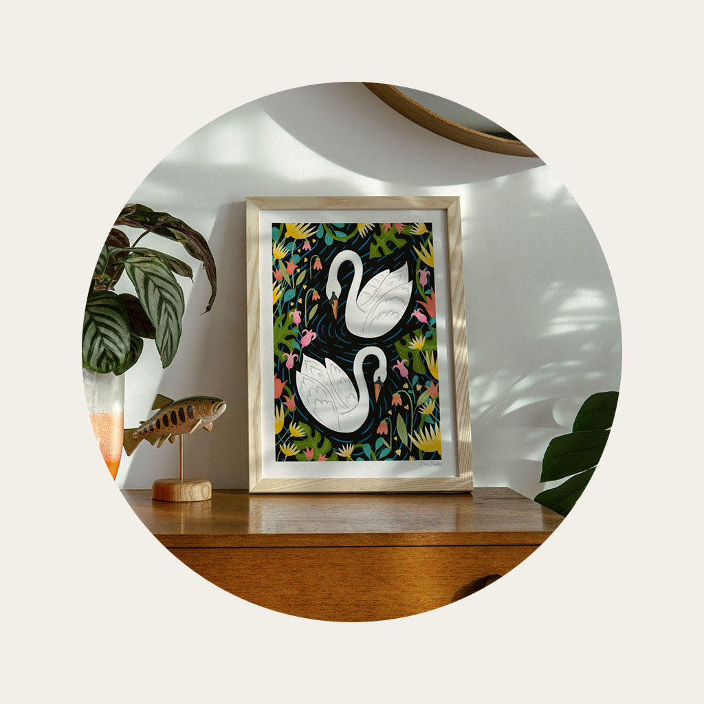 An illustrated art print of two swans is leant against the wall on top of a cabinet, surrounded by house plants.