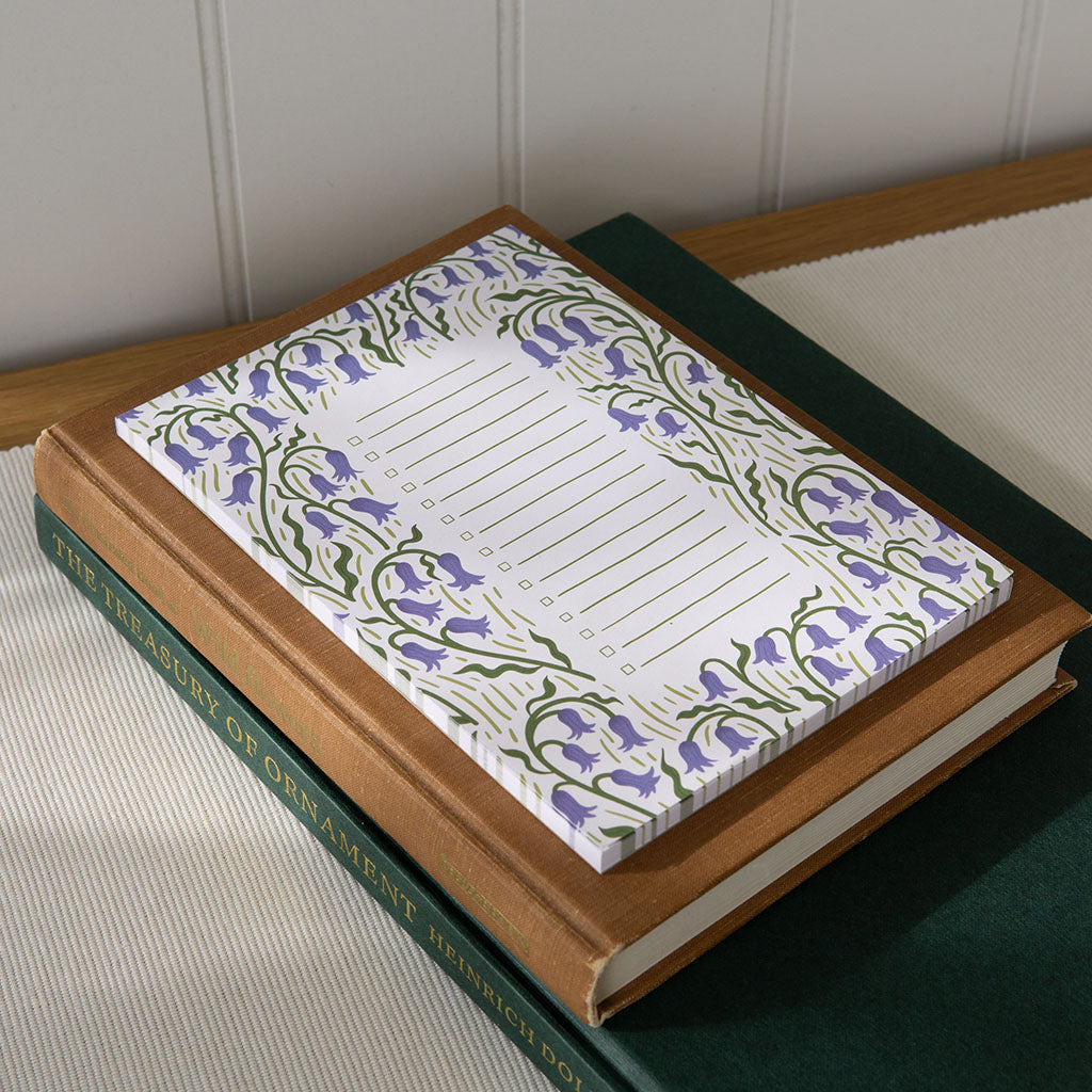 The Bluebells Floral Notepad is a rectangular illustrated checklist pad with a green and purple floral border, perfect for shopping lists or daily tasks, featuring a centred checklist and blank lines on a white background.