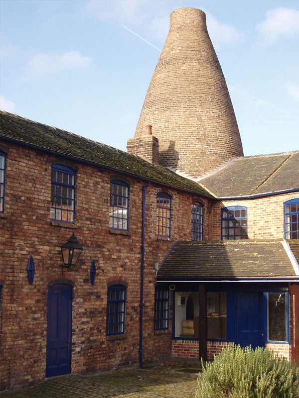 A historic brick building with blue doors and windows stands in front of a large, conical brick kiln under a partly cloudy sky.