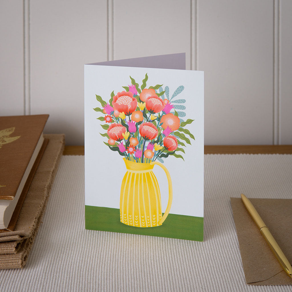 A greetings card with an illustrated vase of flowers on the front sitting displayed on a table.