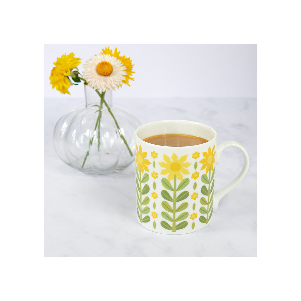A ceramic mug with a yellow floral pattern filled with coffee sits in front of a glass vase holding three yellow and white flowers on a white surface.