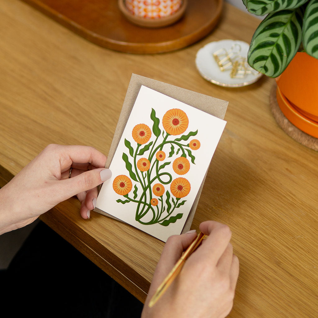 The Pot Marigold Greetings Card stands upright and features an illustration of orange flowers with green stems on a white background, printed on sustainably sourced card-stock.