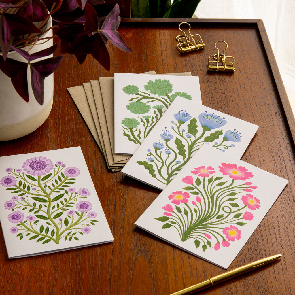 The Folk Flowers Greetings Card Set includes four unique botanical art cards: blue, pink, green, and purple flowers are illustrated in a vintage floral art style, with brown kraft paper envelopes.
