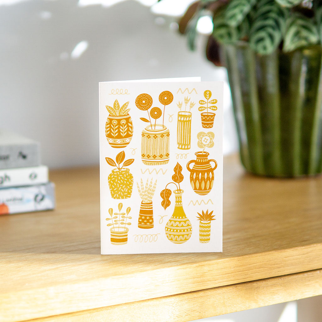 The Yellow Pots & Plants Greetings Card features illustrated yellow and orange cacti and succulents in pots and vases, arranged decoratively on a cream background—ideal for plant lovers.
