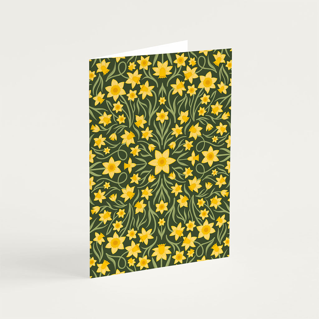The Heritage Daffodils Greetings Card displays a symmetrical pattern of yellow daffodils and green leaves on a dark background, standing upright against a plain surface.