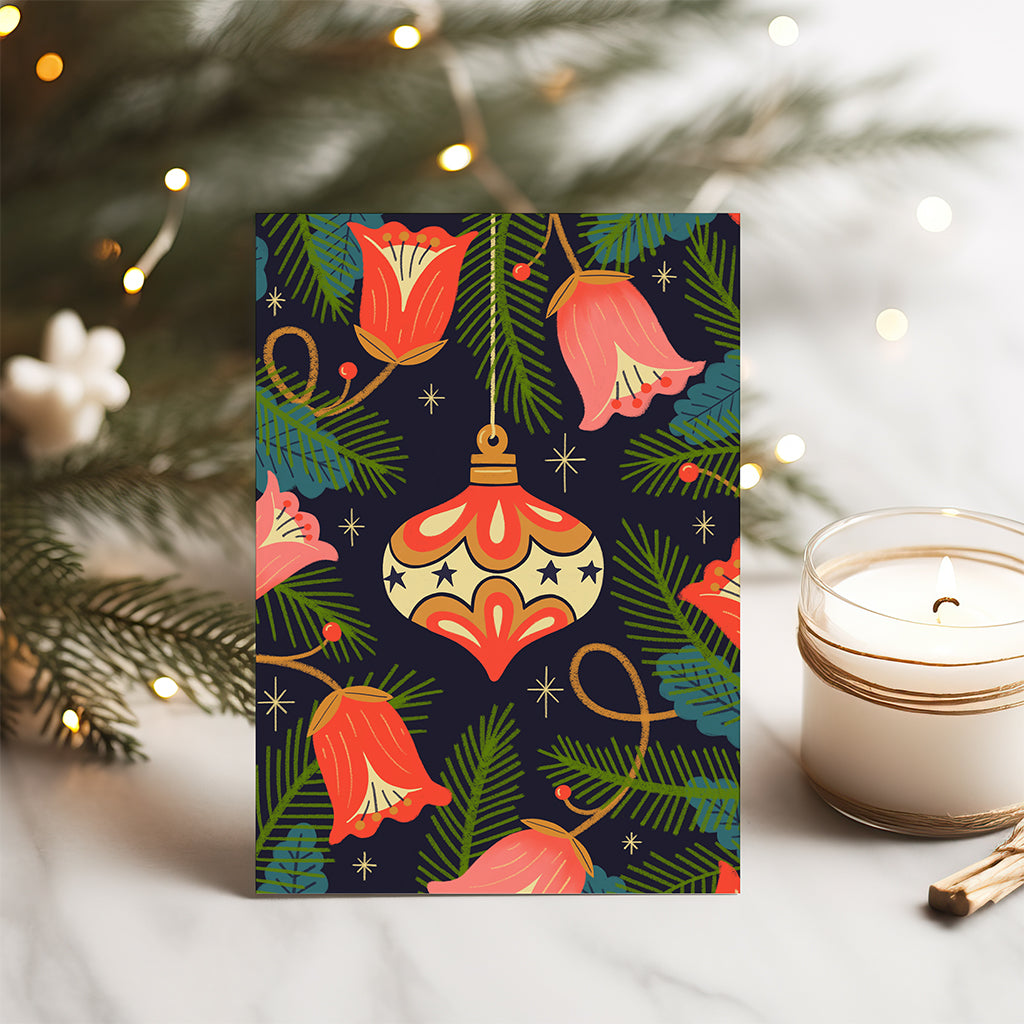 The Bauble Christmas Card features red flowers, green pine branches, gold stars, and a gold ornament on a dark background for festive holiday style.