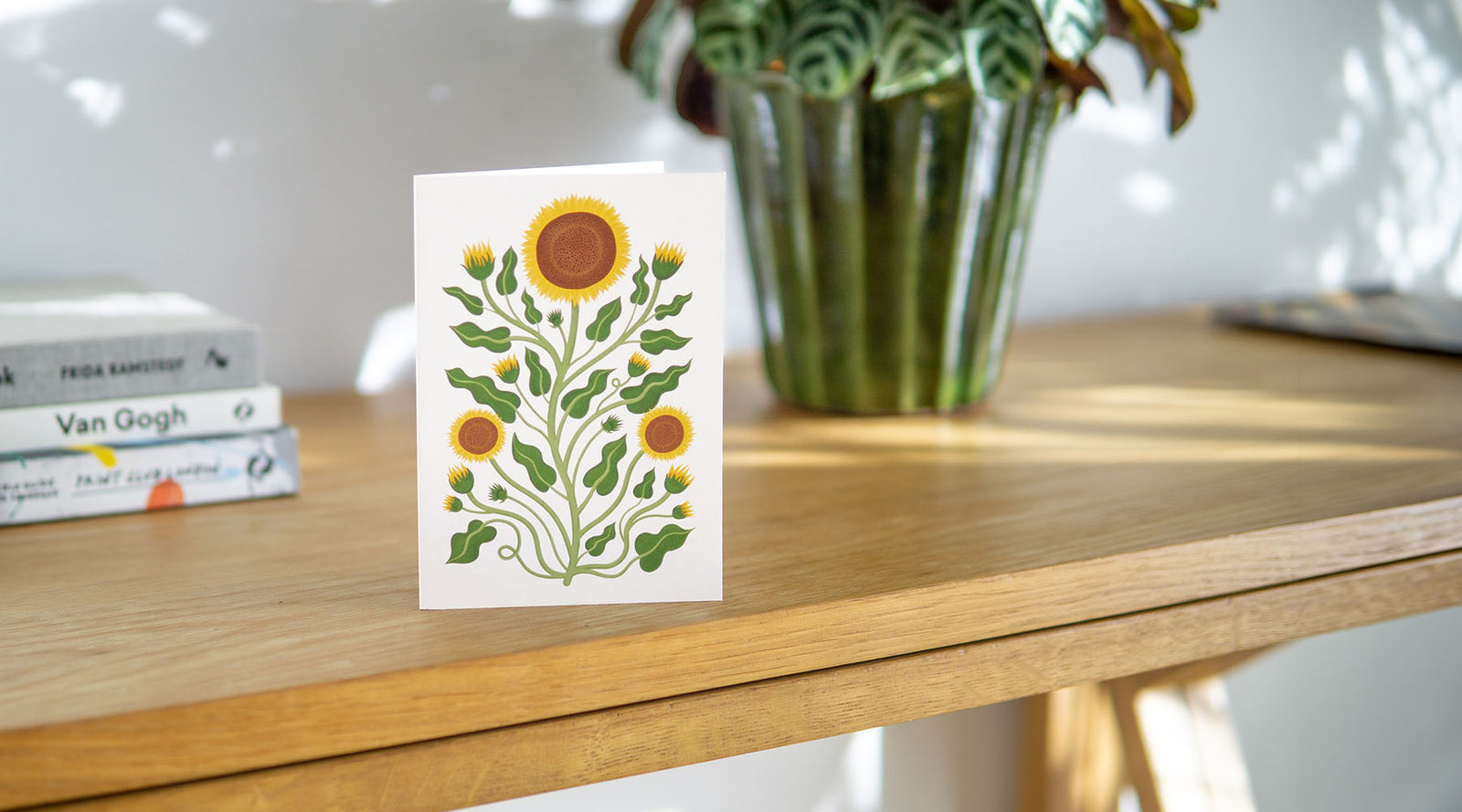A botanical art greetings card with a vintage sunflower design on a table with art books and house plants.