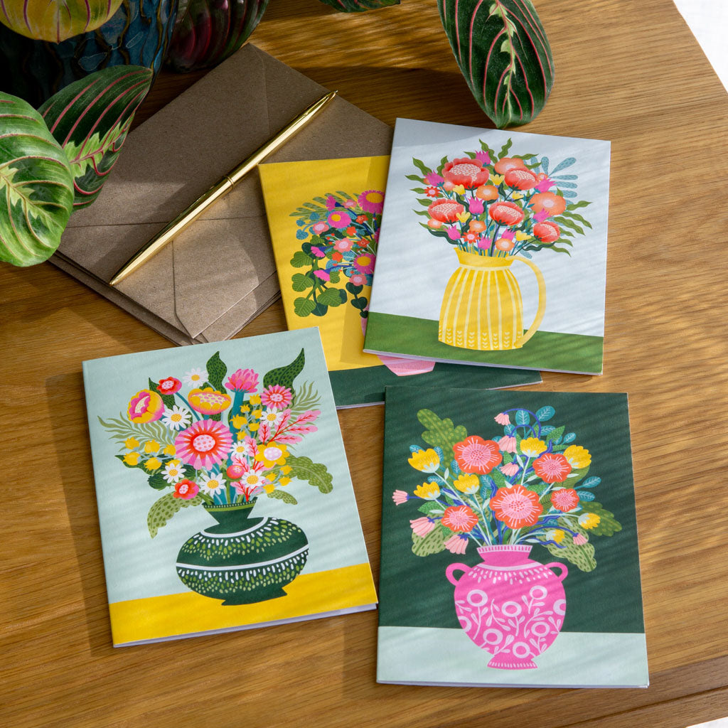 The Flowers in Vases Greetings Card Set includes four cards featuring colourful vintage vases arranged on a wooden table with a leafy plant, envelopes, and a gold pen.