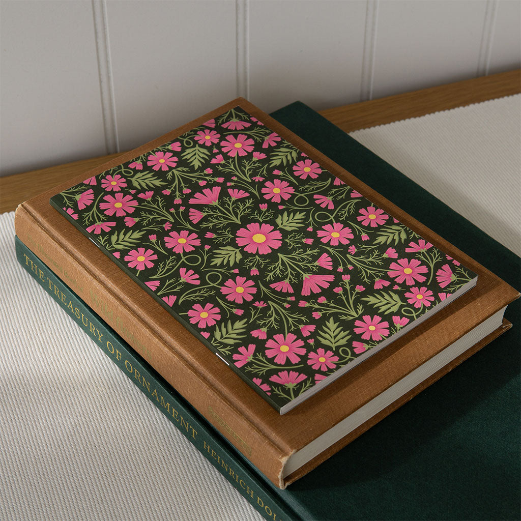 The Heritage Cosmos Notebook is an A5 eco-friendly notebook with a symmetrical floral design of pink flowers and green leaves on a dark cover, crafted from recycled paper for sustainable note-taking.