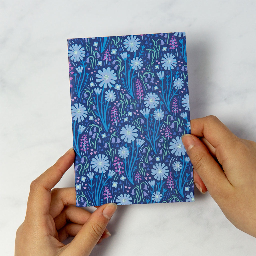 The Navy Blue Wildflowers Greetings Card stands upright with a navy blue background, decorated with white daisies and small purple wildflowers for an elegant floral touch.