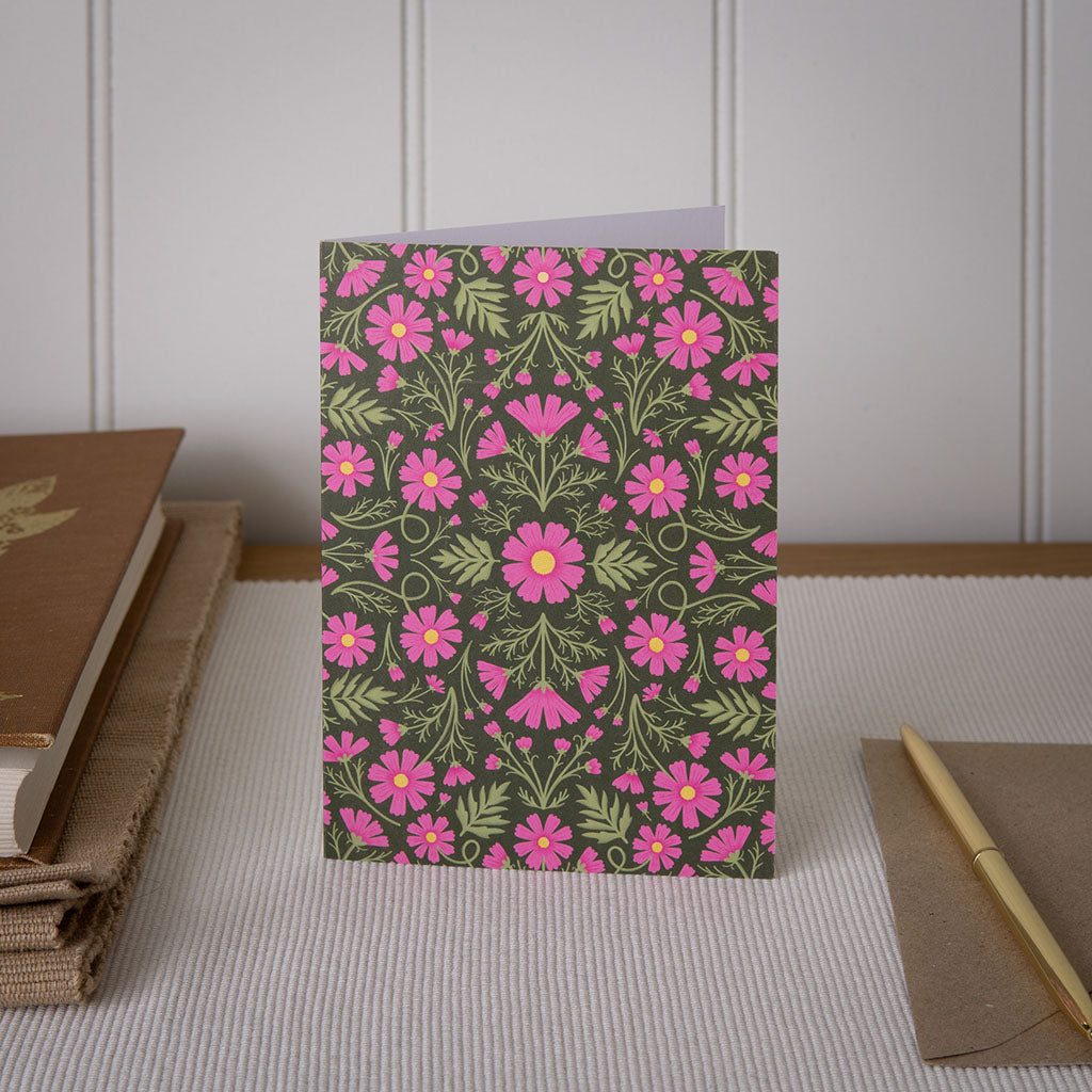The Heritage Cosmos Greetings Card stands upright, showcasing pink flowers and green leaves on a dark background, printed with vegetable-based inks.