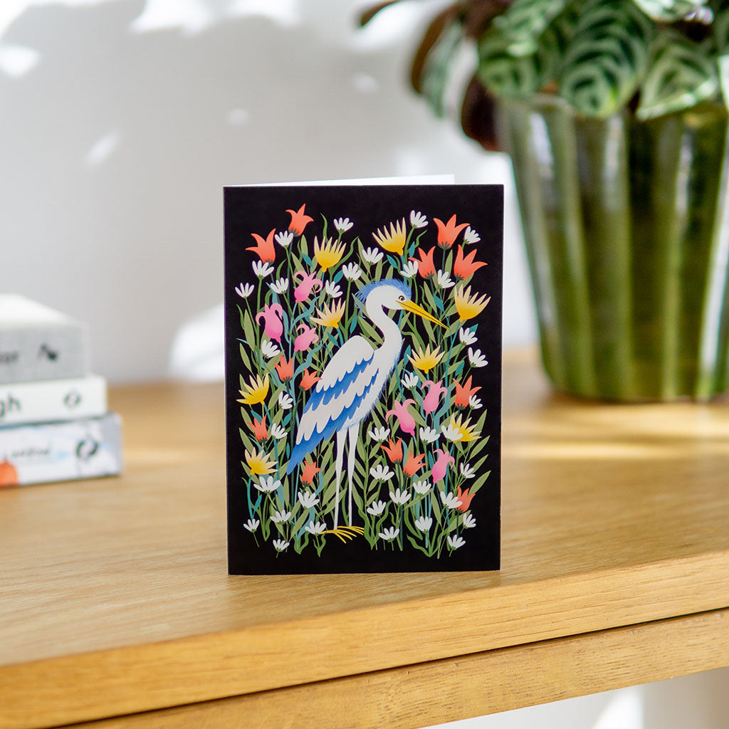 The Heron Greetings Card features a white and blue heron standing among colourful flowers on a black background—ideal for birthdays or as a thoughtful Mothers Day card.