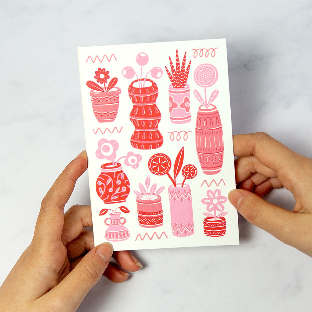 The Red Pots & Plants Greetings Card features abstract red and pink vases with flowers on a light background, an ideal choice for plant lovers.