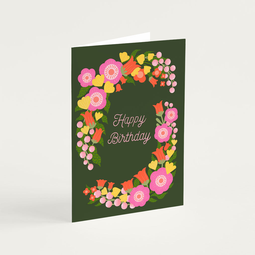 The Happy Birthday Greetings Card features a dark green background with Happy Birthday in the center, encircled by a vibrant floral wreath of pink, yellow, and orange flowers, a perfect choice for any birthday celebration.