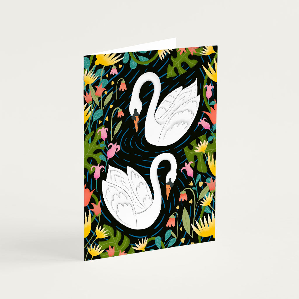 Swans Greetings Card featuring two white swans swimming among colorful illustrated flowers and green foliage on a dark background. Perfect for anniversaries and special occasions.