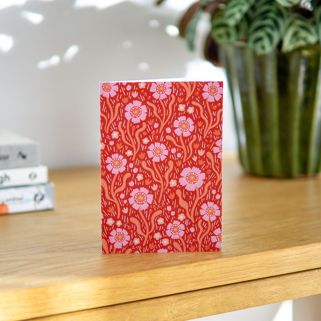 The Red Wildflowers Greetings Card stands upright, showcasing a red background with pink and orange dog roses and leaves inspired by the beauty of British wildflowers.