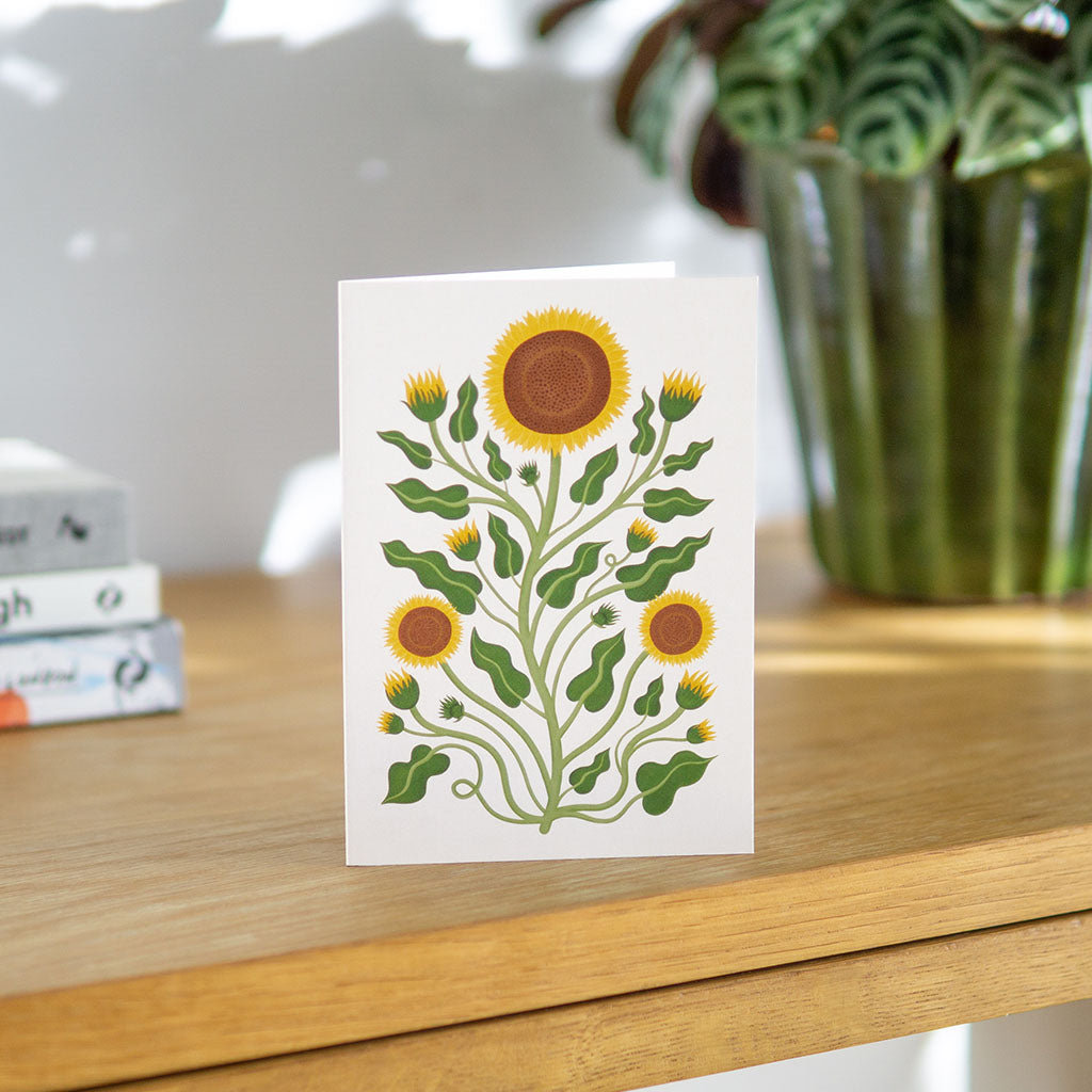 The Sunflowers Greetings Card stands upright on a wooden table, with a green potted plant and stacked books behind it—ideal for birthdays or Mother’s Day.