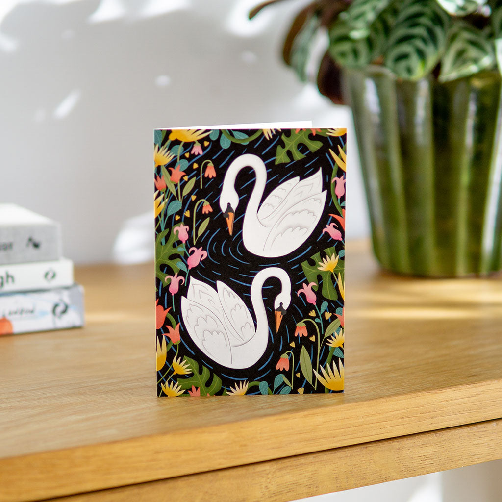 Swans Greetings Card featuring two white swans swimming among colorful illustrated flowers and green foliage on a dark background. Perfect for anniversaries and special occasions.