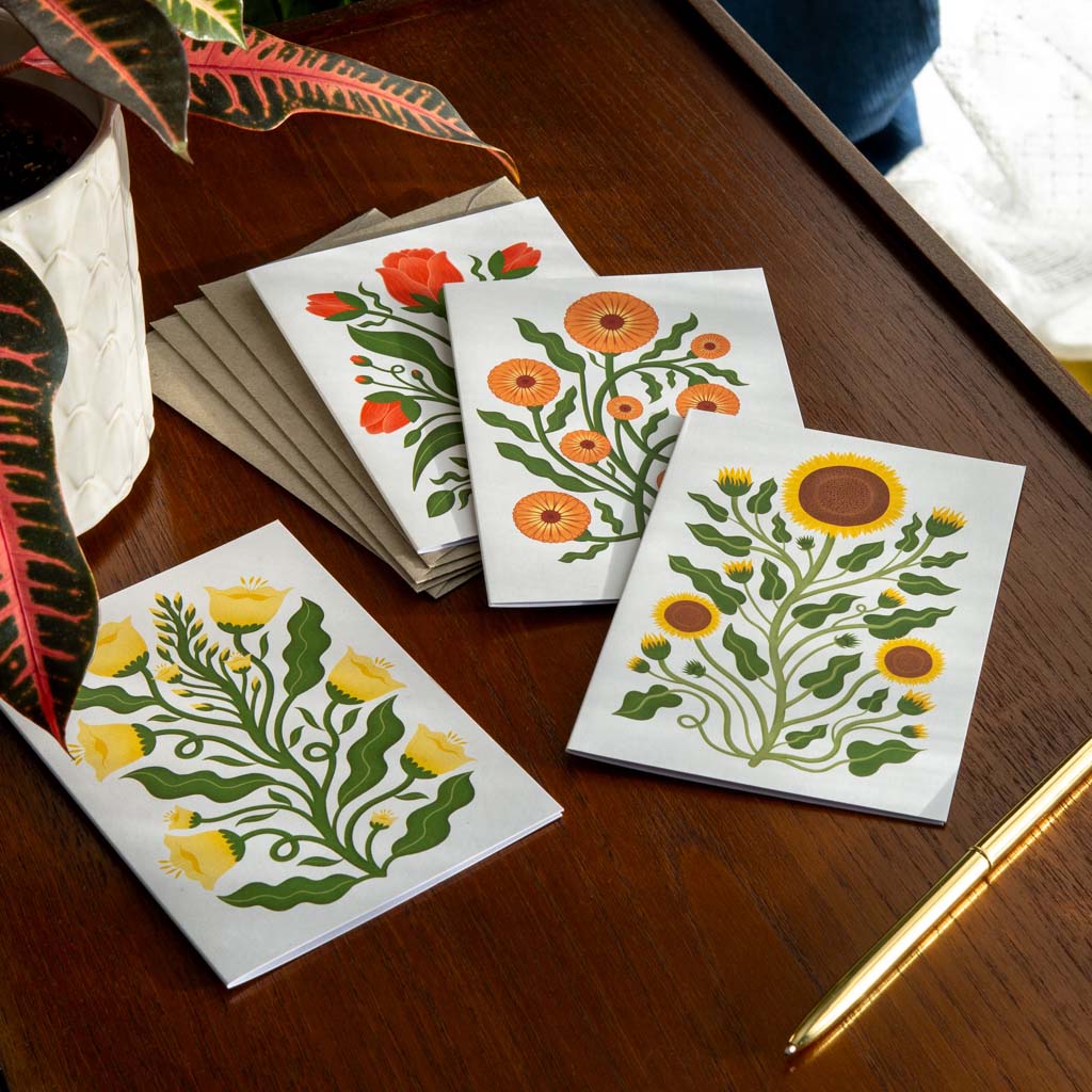 The Folk Flowers Greetings Card Set, featuring four illustrated cards with botanical art including a sunflower design, is displayed on a wooden table with a pen, envelopes, and a potted plant.