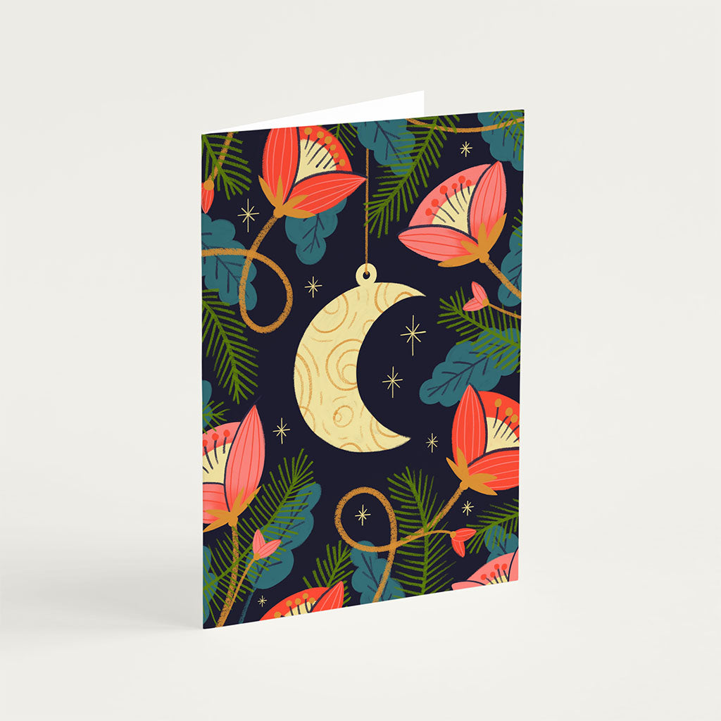 The Moon Christmas Card features a gold crescent moon pendant surrounded by pink flowers, green leaves, and small stars on a dark background, adding holiday magic.