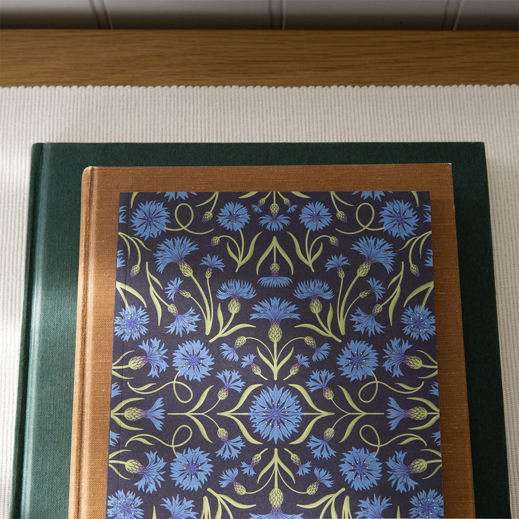 The Heritage Cornflowers Notebook features a vintage-inspired dark cover with a symmetrical pattern of blue cornflowers and green stems.