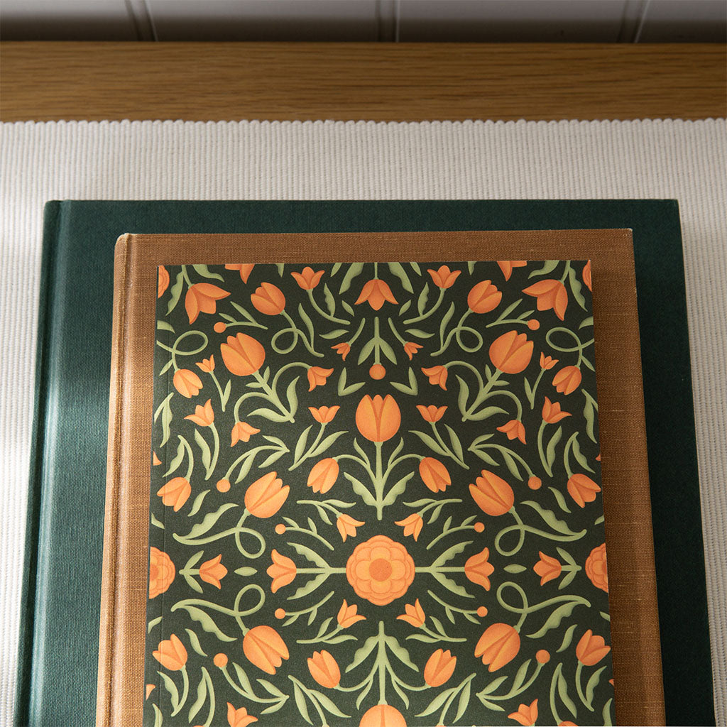 The Heritage Tulips Notebook, with its vintage-inspired cover of orange and green tulips on a dark background, is displayed standing against a plain white surface.