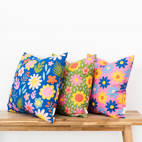 Three decorative pillows with colorful, bold floral patterns in blue, green, and pink tones are arranged on a wooden bench against a plain white background.