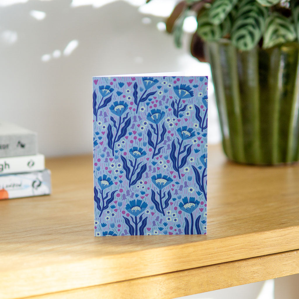The Blue Wildflowers Greetings Card features a blue botanical pattern inspired by British wildflowers on a light purple background, with illustrated flowers and leaves.