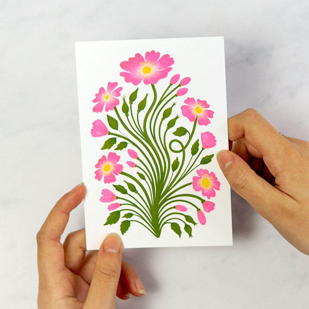 The Wild Roses Greetings Card stands upright and features an illustration of pink wild roses with green stems and leaves on a pale cream background.