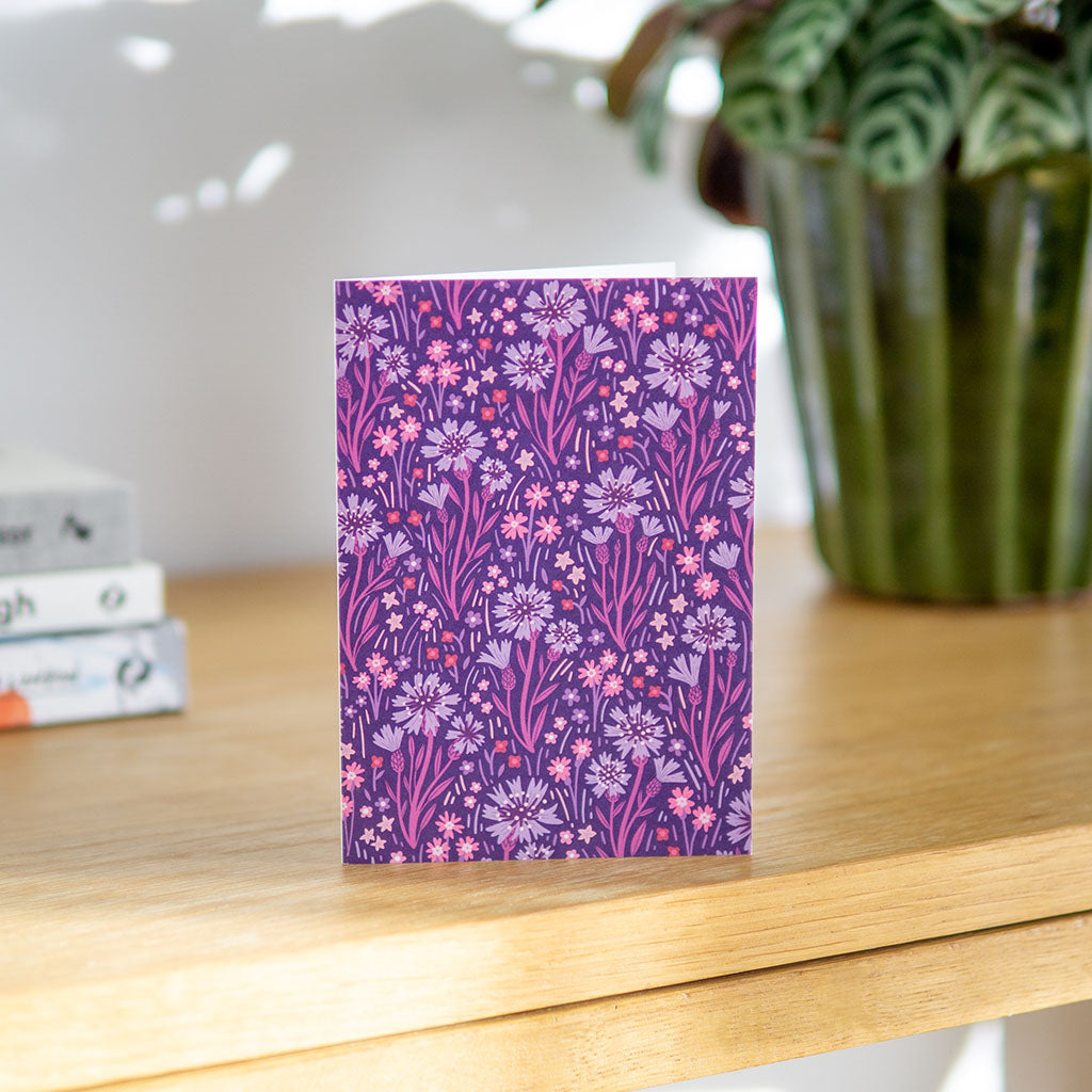 The Indigo Wildflowers Greetings Card stands upright, showcasing a decorative purple background with pink, lavender, and indigo wildflowers and foliage.