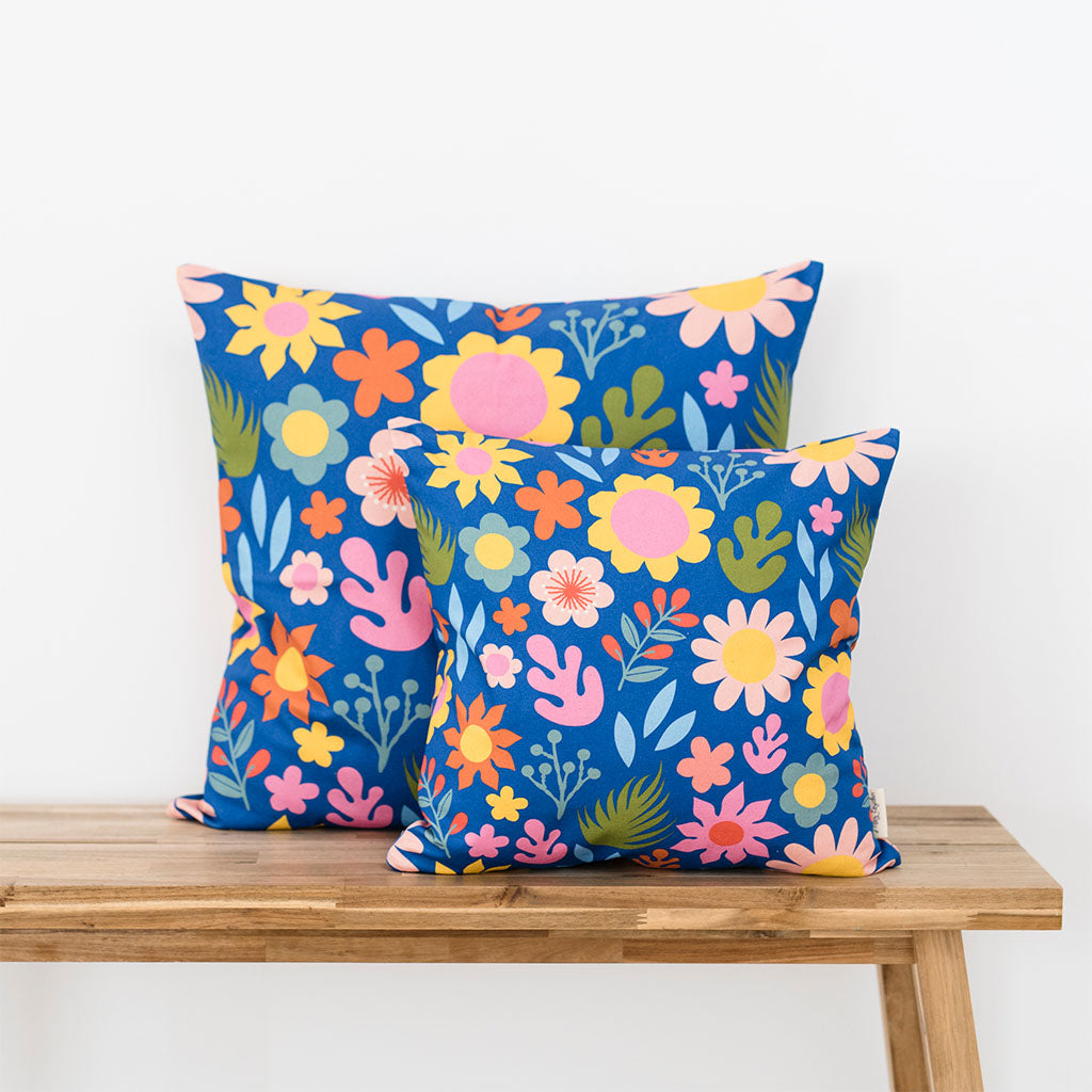 Two square throw pillows with Flower Patch Cushion Covers, featuring vintage floral patterns on a blue background and made from organic cotton, sit on a wooden bench against a white wall for added comfort.