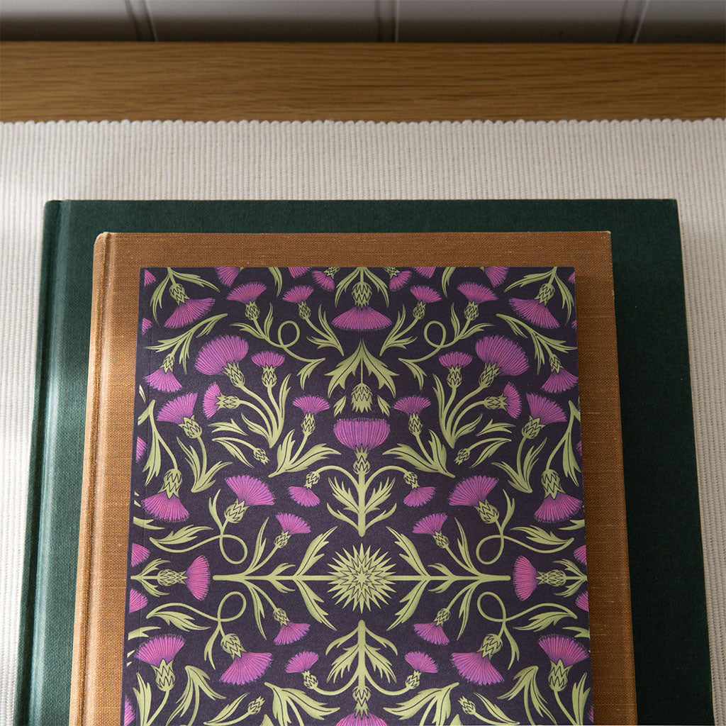 The Heritage Thistles Notebook features lush, symmetrical purple thistle flowers and green stems on a dark background, offering a vintage-inspired floral design ideal for journaling enthusiasts.
