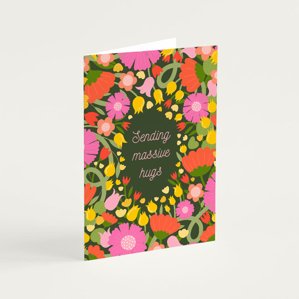 The Massive Hugs Greetings Card is sustainably made and features pink, red, and yellow flowers on a dark green background with Sending massive hugs in cursive at the center.