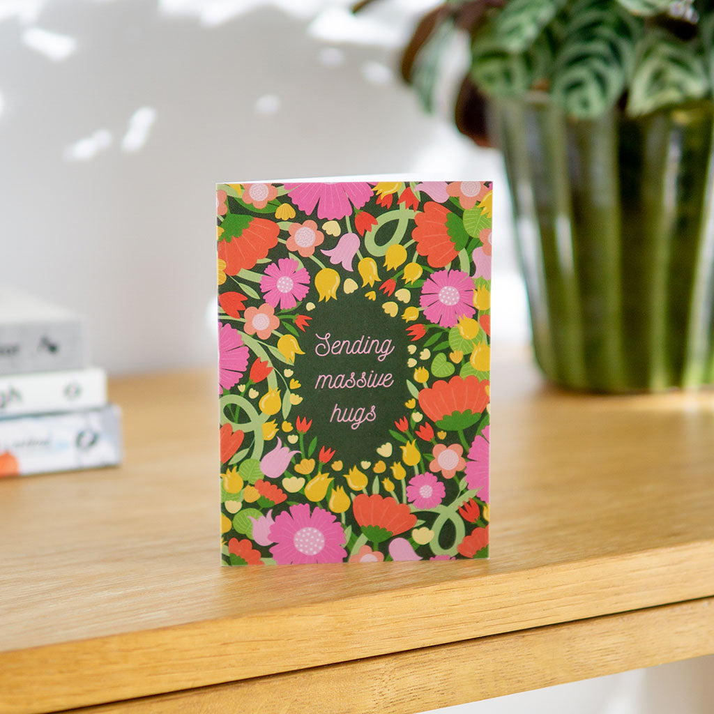 The Massive Hugs Greetings Card is sustainably made and features pink, red, and yellow flowers on a dark green background with Sending massive hugs in cursive at the center.