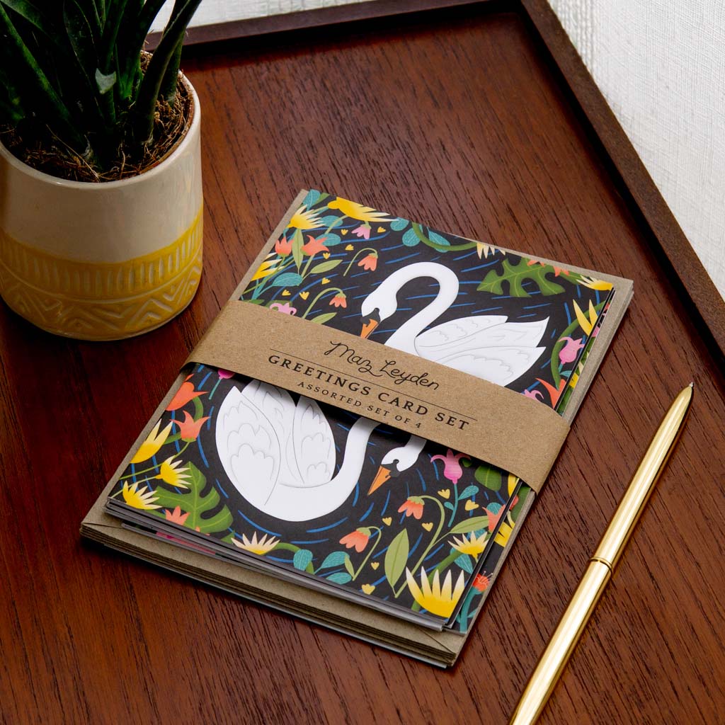 The British Birds Greetings Card Set, featuring a white swan design, rests on a wooden table alongside matching envelopes, a gold pen, and a potted plant.