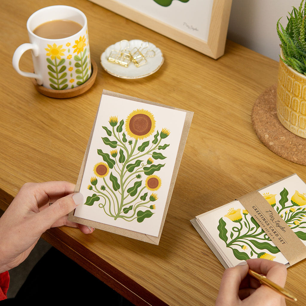 Someone writes on the Sunflowers Greetings Card at a wooden desk with tea, paper clips, and a potted plant—an ideal choice for sending cheerful birthday wishes or thoughtful messages for Mother’s Day.