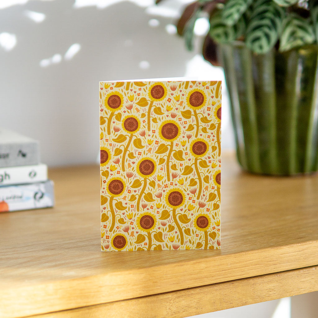 The Yellow Wildflowers Greetings Card stands upright and features a repeating pattern of sunflowers and yellow wildflowers on a light background.
