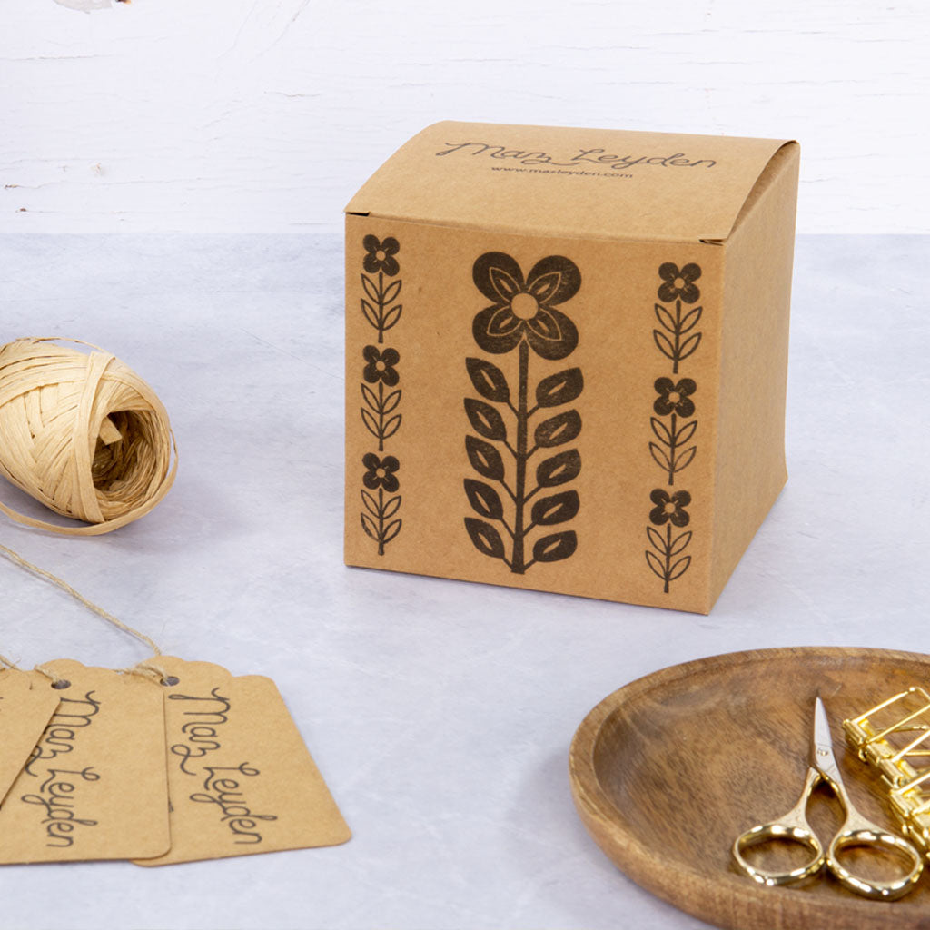 A brown cardboard box with a black floral design sits on a table next to a ball of twine, gift tags, a wooden plate, gold scissors, and binder clips.