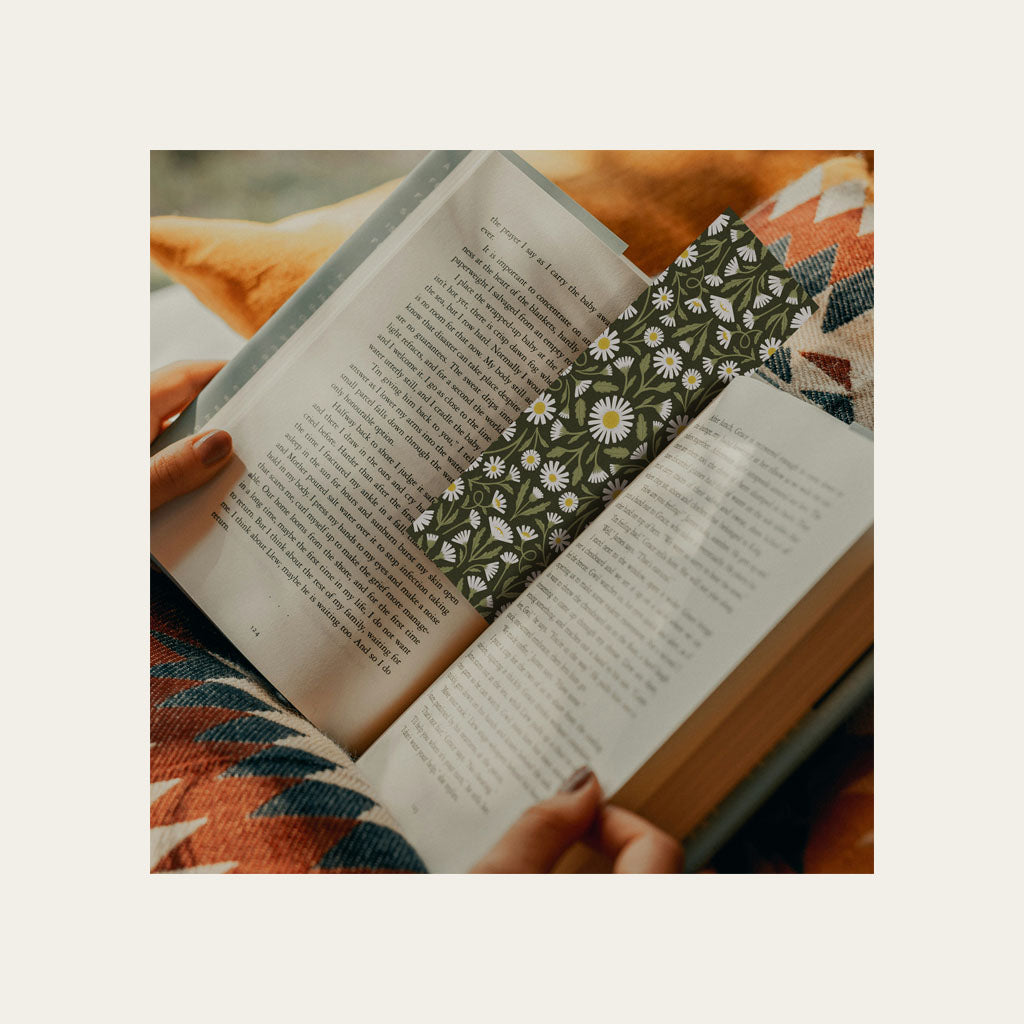 A person holds an open book with a green floral bookmark on patterned fabric.