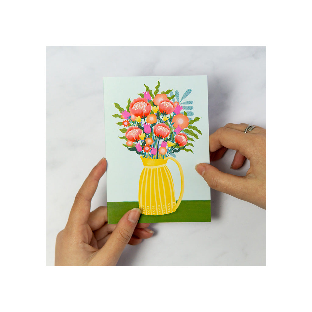 A person holds a greeting card featuring an illustration of a yellow vase with pink flowers on a white background.