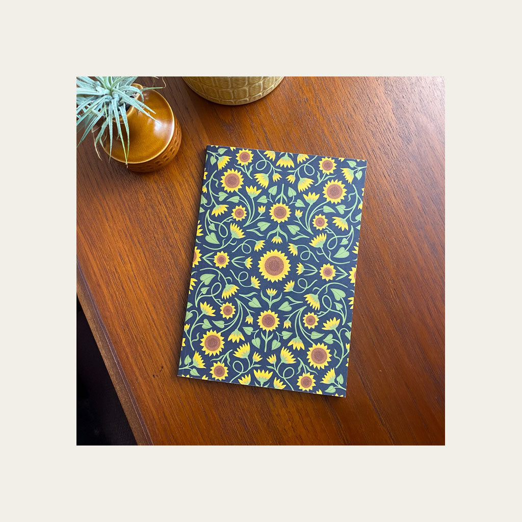 A notebook with a yellow sunflower pattern on a dark background sits on a wooden table next to a small plant and a textured container.