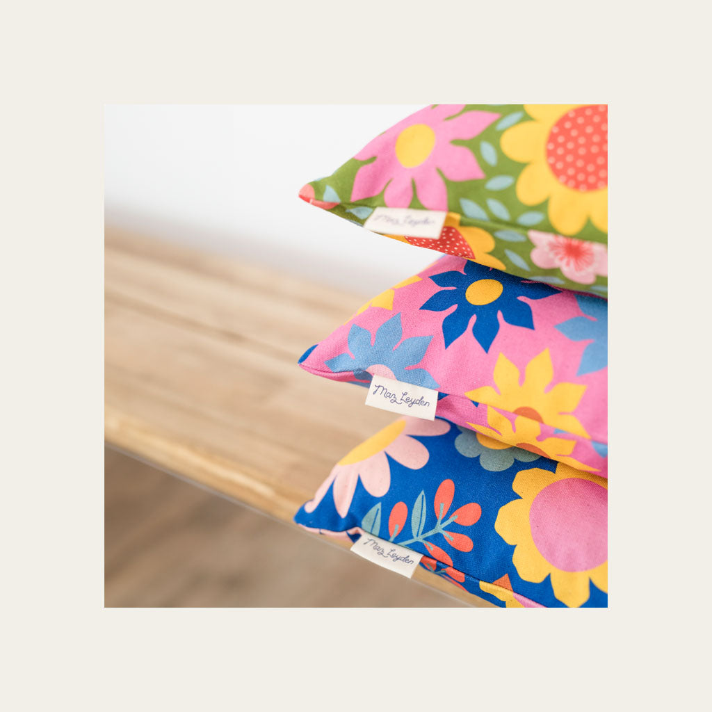 Three colorful floral-patterned pillows with visible fabric tags are stacked on a wooden surface against a neutral background.